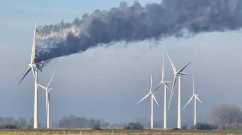 Rob Ambrose Smoke is billowing from a wind turbine that is on fire in a field. There are five other turbines nearby. The sky is blue and there is a field in the foreground and trees in the background