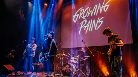 Four young musicians are on stage playing instruments. A large sign reads: 'Growing Pains' in the background. 