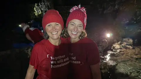 Two women in red woolly hats and smiling with their arms around each other. They are both wearing red t-shorts which says: “Together we will end homelessness.”