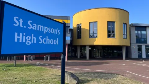 A blue sign saying St Sampson's High School with a yellow cylindrical building in the background, held up with grey stilts. 