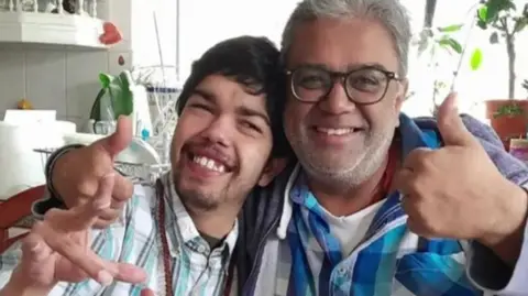 Balram Patel and his father Jay Patel smile at the camera. Jay Patel is showing a thumbs up symbol.