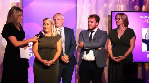 The team of In Charley's Memory are standing on stage next to a presenter who is holding a microphone. They are dressed in suits and dresses. In the background is the purple branding of BBC Radio Somerset.