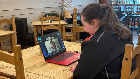 A girl sits in a cosy cafe, she is looking at a laptop which is open on the table in front of her. The laptop has an image of a woman on the screen in the middle of a video call. The girl is wearing a black and red uniform and has long dark hair pulled back in a ponytail. 