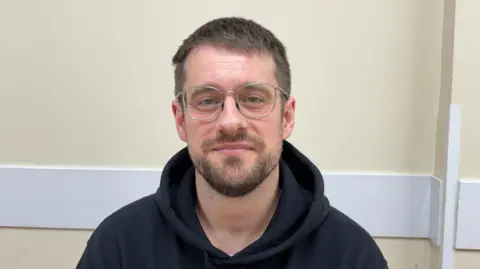 Photograph of Hughes who is looking straight into the camera and smiling slightly. He has short brown hair and a short beard, and he is wearing silver-framed glasses and a black hoodie. He's sat indoors, in a room with cream painted walls. 