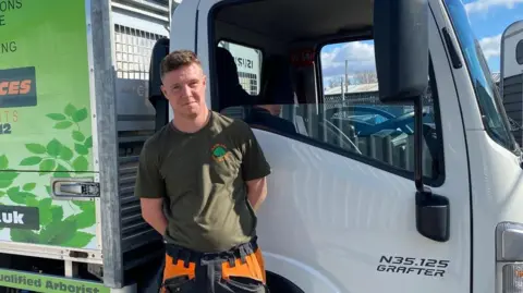 Jacob Francis standing in front of the driver's door of a large white van. His hands are behind his back. His hair is short and brown and he is wearing a khaki t-shirt.
