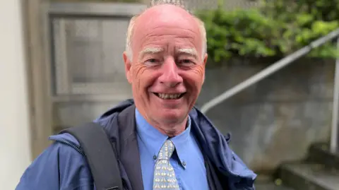 BBC Deputy George Oswald - A man with a bald head and white hair on the side of his head. He is wearing a blue coat, blue buttoned up shirt and a pastel blue and white tie. 