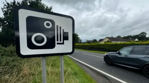 A speed camera sign at the side of a road where a black car is driving past.