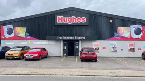 Exterior of a Hughes shopfront. The fascia of the front has images of a washing machine, TV screen and a laptop. Cars are parked outside the store
