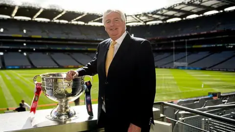 Getty Images A man with short grey hair stands with his right arm resting on a large silver bowl trophy, with red and navy ribbons around the trophy's handles. Behind him is a large empty stadium with a large green pitch in the centre. The man is wearing a black suit, white shirt and yellow tie. 
