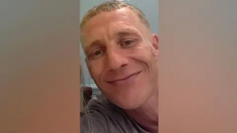 Humberside Police Shane Boxall has short-cropped blond hair and hazel eyes. He is wearing a grey T-shirt and is smiling into the camera.