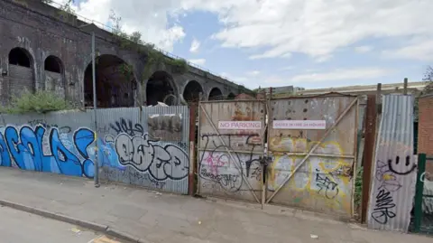 Metal fences covered in graffiti and a metal gate with no parking notice on it with a viaduct in the background