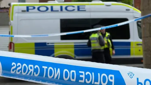 Seen behind a police cordon, two unformed officers stand beside a police van, talking