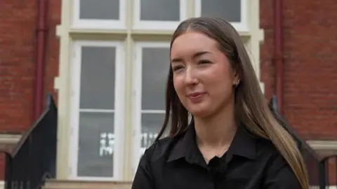 Richard Knights/BBC A young woman with long brown hair looks to the left of the camera. There is a red brick building with a window in the background.