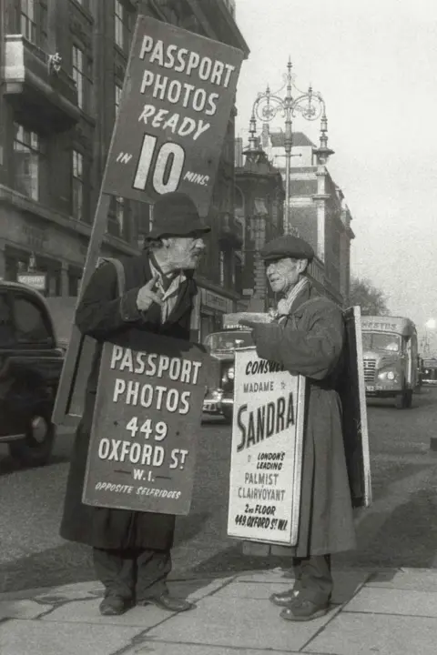 Philip Sharkey Black‑and‑white street photograph showing two people standing on a pavement holding advertising boards. The boards promote passport photos ready in ten minutes and display an Oxford Street address. Buses and buildings line the street in the background.