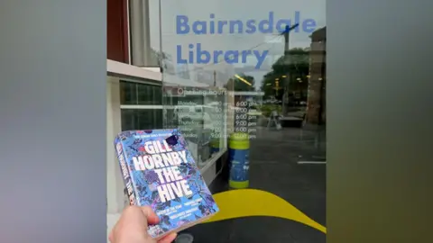 Bairnsdale Library A hand holds a blue book titled Gill Hornby The Hive is heldnext to a door with opening times, the words Bairnsdale Library is on the door