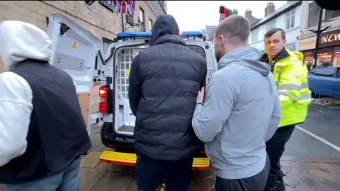 Shaun Whitmore/BBC A combination of plain-clothed and uniformed police officers take an arrested suspect into the back of a police van.
