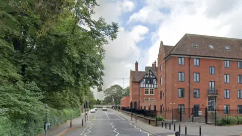 Google A single carriageway road with green trees to the left and tall brick buildings on the right