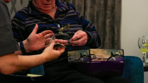 Corgi A man in a multi-coloured top is holding a Spitfire toy model. He is sitting next to another man who is also holding a part of the Spitfire model. 