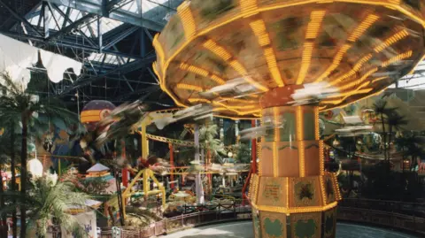 Metrocentre Fairgroung swings which is in motion. People on the swings are blurred and the centre of the ride is tilted and lit up. There is a yellow rollercoaster behind the swings and other rides on the floor below. The room is covered in palm trees and the ceiling is largel glass with beams. 