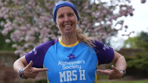 Apryl wearing a light blue running vest with Alzheimer's Society written on the front - the charity she's running in aid of at Sunday's London Marathon. Her top also says Mrs H. She's smiling and pointing to her name on her vest.