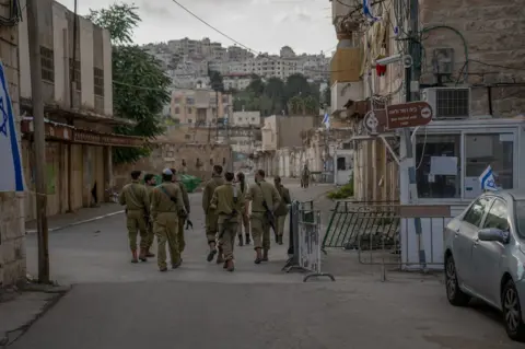 BBC Israeli soldiers walk past Fawaz Qafisha's house. The Palestinian residents are not allowed on the street.