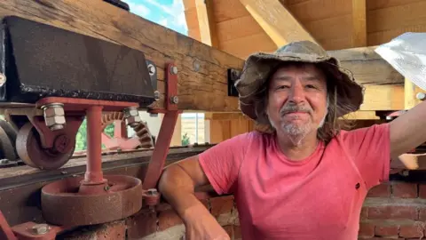 Andrew Turner/BBC Paul Kemp standing inside the cap of the wind pump, wearing a pink tee shirt and wax hat. He has a greying beard and has one arm resting on the brickwork.