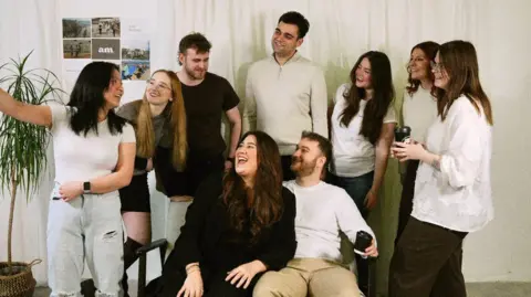 Adela Rokytova A group of nine people gathered to take a photo. There is a man and a woman sitting on a bench, and there are five women and two men standing behind them. One woman has her arm raised taking a selfie of the group, but the phone is not visible. They are all smiling.