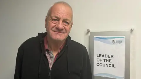 LDRS Milan Radulovic wearing a black hoodie and red collared shirt in front of a sign which says leader of the council