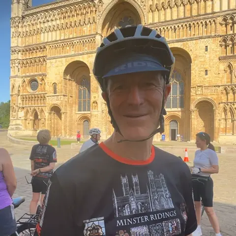 Callum Davis / BBC A head and shoulders shot of Kev Stroud wearing a cycling helmet and a T-shirt with a drawing of Lincoln Minster and York Minster on. It also reads"MINSTER RIDINGS" in white capital letters. He is smiling at the camera and is stood in front of Lincoln Minster. Other cyclists are stood around him.