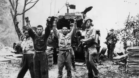 Universal History Archive/Universal Images Group via Getty Images Three Korean men in military clothing hold their hands up, with what appears to be a US solider with a gun standing behind them. His helmet is sitting at an angle on his head. Behind them is a tank, and the outline of more soldiers can be made out. The picture is black and white. 