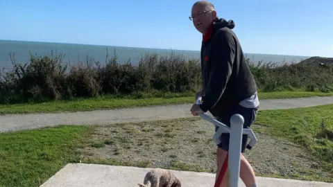 Mark Phillips A man with greying hair and glasses wearing a black hoodie and navy shorts on outdoor gym equipment next to a coastal path. The man is off-centre and in side profile, his head turned to the camera. A small dog's back can just be seen at his feet. In the background is grass, a gravel path, hedgerow and, beyond it, the sea.