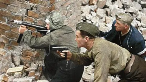 An old photograph, with colour added later, of three Polish resistance fighters. Wanda is on the far left. They are crouched behind a wall aiming a gun. The ground is covered in rubble.