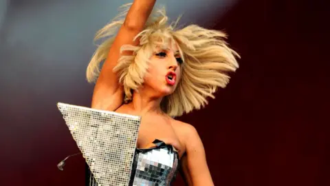 Lady Gaga on stage with one arm in the air in an elaborate mirrored dress at Glastonbury in 2009