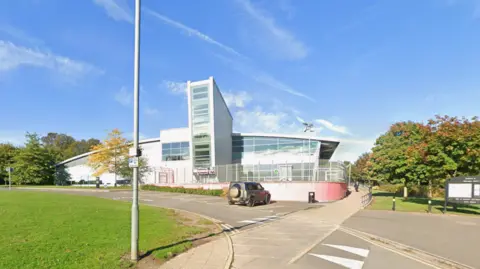 A building that looks white in the sun on this photo is in the background and includes a large rectangular-shaped section in the middle of the visible side on the image. Grass is on the left, trees are on the left and right and a road goes from the left to the bottom right of the photo. 