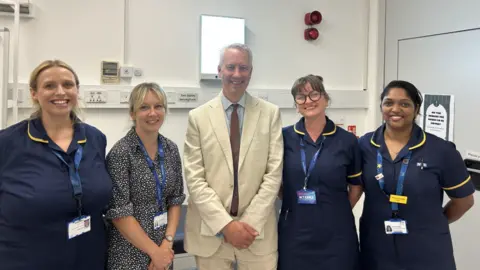 Somerset NHS Foundation Trust Five clinicians - four women with a man in the middle - are all smiling to camera in a hospital-type office. The man is wearing a smart beige linen suit and brown tie and three women are in navy blue nurse uniform and wearing lanyards. The other woman is in a black and white dress.