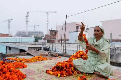 Reuters Shakila Bano, 38 years old, makes a garland of Marigold flowers at her house as the construction site of Hindu Ram Temple is seen in the background, in Ayodhya, India, November 22, 2023. REUTERS/Anushree Fadnavis