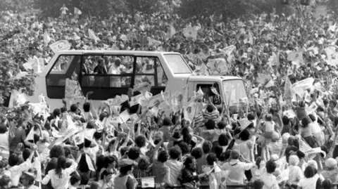 PA Media A black and white photo of Pope John Paul II framed in the Popemobile, surrounded by thousands of people, on his arrival at Bellahouston Park, Glasgow for a Mass