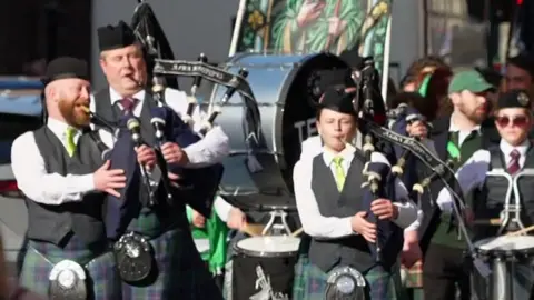 Iris marching band with pipes, drums, dark forage caps, light green ties, white shirts and green tartan kilts.