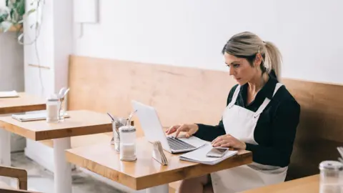 Getty Images Female cafe manager wearing a black shirt and white apron, sitting at cafe table looking at her laptop