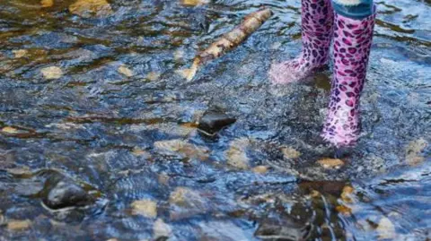 Water running over rocks in a shallow river with someone standing in the water wearing brightly-coloured wellies.