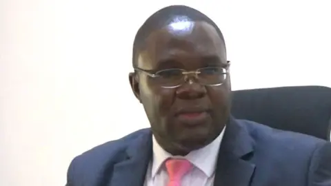 The magistrate in a dark jacket and pint tie looks at a laptop computer to his left.  