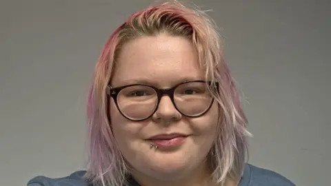 Supplied A head shot of a young woman with light pink shoulder-length hair and round black-rimmed glasses. The woman has two nose rings and piercings on her mouth. She is smiling directly at the camera.