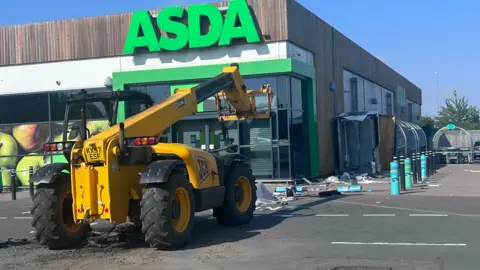 Telehandler outside a branch of Asda - a large green-coloured Asda logo is on the store front. Debris from smashed bollards is strewn in front of the telehandler. 