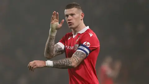 Pa Media James McClean wearing the Red Football Shirt of Wrexham during a match. He has short brown hair and tattoos on both arms he wears the captains Armband and raises his right arm raised 