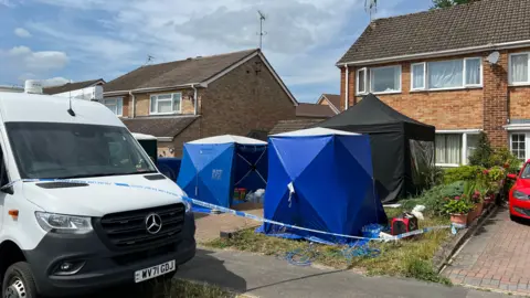 BBC Three tents in the front garden of a semi-detached house in Sandhurst. A white van is parked on the side of the road and police tape surrounds the front of the house and the driveway.