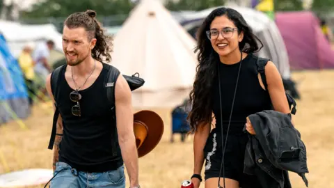 @garryjonesphotography A man and a woman smile as they walk through the WOMAD festival site in Wiltshire. The man is wearing a black vest top, has long hair and is carrying a rucksack. The woman has long black hair, is wearing glasses and also carrying a rucksack. The grass behind them is light brown in colour