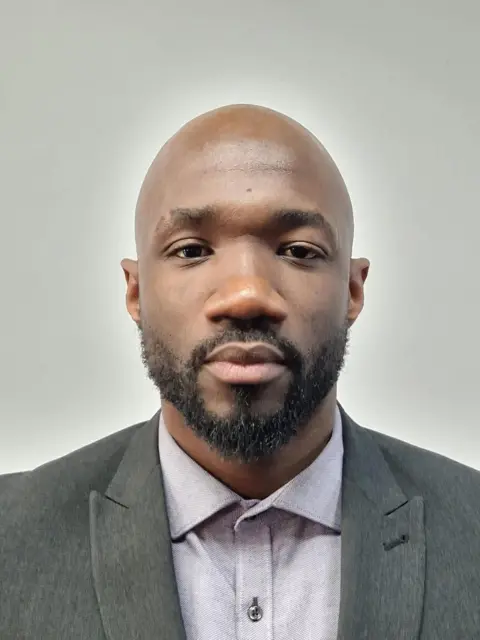 A man with a beard looks directly into the camera. He is wearing a grey blazer and a buttoned up light grey shirt.