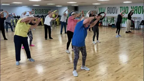 La instructora de baile y fitness Maureen Conroy imparte clases en un gran gimnasio. Mantiene las manos por encima de la cabeza y sus alumnos siguen sus movimientos detrás de ella. El piso del gimnasio es de madera pulida.