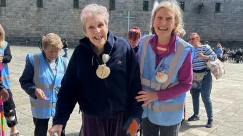 An elderly lady smiles broadly at the camera. She is leaning on a walking stick and a wheelchair can be seen just behind her. Next to her is a younger woman, also smiling into the camera.