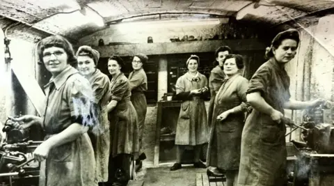 A group of women work in a makeshift munitions factory beneath the arcade in New Brighton, Merseyside. They are dressed in work overcoats and have their hair tied up or under headscarves as they operate machinery making bullets and shell casings. 